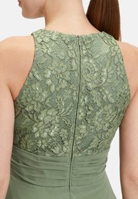 Robe verte avec un dos en dentelle et des motifs floraux, un corsage en tissu lisse et une fermeture éclair dissimulée dans le dos. Design texturé et superposé.