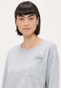 Sivý bavlnený sveter s okrúhlym výstrihom, dlhými rukávmi a kvetinovým dizajnom na ľavej náprsnej vrecku. Mäkka textúra.