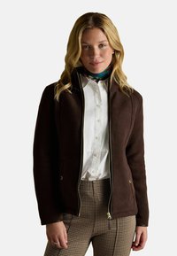 Blondynka w ciemno-brązowej kurtce na zamek, nosząca białą bluzkę z guzikami i brązowe spodnie w wzór hounds-tooth, stojąca na jednolitym tle.