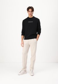 Fekete kötött pulóver bordázott mandzsettákkal, a "GUESS JEANS" logóval. Világos bézs nadrág és fehér sportcipő. Egyszerű, testhezálló fazon.