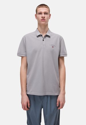 Graues Poloshirt mit weicher Textur, Drei-Knopf-Leiste, kurzen Ärmeln und kleinem Logo-Detail auf der linken Brust. Getragen mit blauen Hosen.