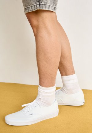 Fehér vászon sportcipők lapos fűzőkkel, alacsony profilúak, és texturált gumitalppal; fehér bordázott zoknikkal viselve, sárga háttér előtt.