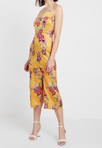 Gelbes Satin-Kleid mit buntem Blumenstickerei, Midi-Länge, verstellbare Träger und einem Schlitz vorne, kombiniert mit weißen Ankle-Strap-Absätzen.