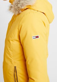 Gele geïsoleerde jas met een met bont gevoerde capuchon, met ritssluitingen afgedekte zakken en een merklogo op de mouw. Glad, duurzaam materiaal.