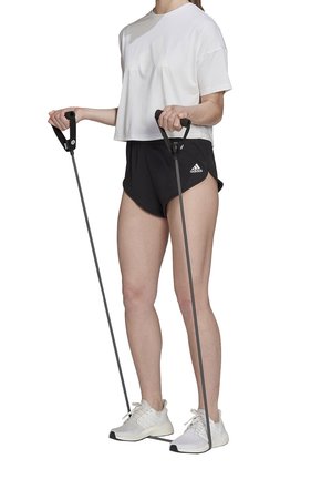 Persoon draagt wit shirt, zwarte Adidas korte broek en witte sneakers, oefent met weerstandsbanden onder de voeten en houdt de handgrepen vast.