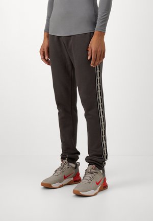 Persoon in donkergrijze sweatpants met merkstrips aan de zijkant en lichtgrijze Nike sneakers met rode accenten, staande op een witte vloer.
