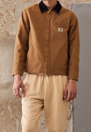 Mężczyzna w brązowej kurtce Carhartt z czarnym kołnierzem i beżowych dresach Nike na tle teksturowanej ściany.