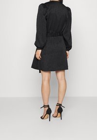 Svart klänning med ett strukturerat mönster, puffärmar och draperad midja. Bärs med svarta högklackade sandaler som avslöjar vristen.