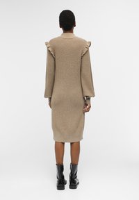 Personne portant une robe en tricot beige midi avec des épaules froncées et de longues manches bouffantes, assortie à des bottes noires à la cheville, se tenant de dos.