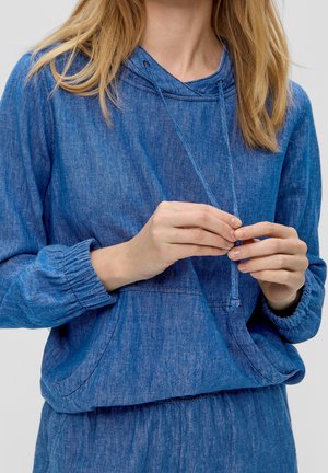 Femme portant un sweat à capuche en jean bleu et un pantalon assorti, ajustant le cordon de la capuche avec ses deux mains.