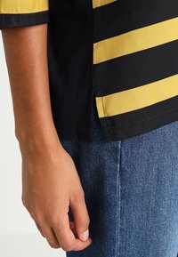 Gestreept shirt met gele en zwarte kleuren, met een gladde textuur en een zijwaarts splitontwerp. Gecombineerd met blauwe spijkerbroeken.