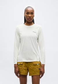 Γυναίκα που στέκεται μπροστά, φοράει λευκό μπλουζάκι μακρυμάνικο της Patagonia και μουσταρδί κίτρινο σορτς cargo, πάνω σε απλό φόντο.