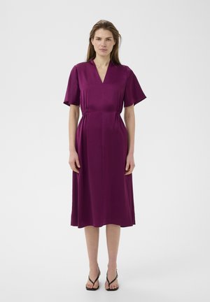 Kobieta stojąca w fioletowej sukience do kolan z krótkimi rękawami, dekoltem w serek, marszczonym stanem i czarnych sandałach na jasnym jednolitym tle.
