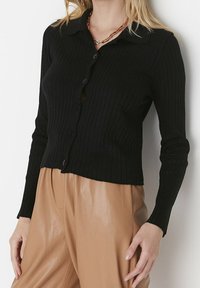 Černý žebrovaný kardigan s dlouhými rukávy a límečkem, se zapínáním na přední straně a zkráceným střihem, v kombinaci s béžovými kalhotami.