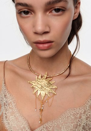 Mujer con blusa de encaje beige y un collar dorado con forma de sol, con cadenas colgantes y pequeñas cuentas que parecen perlas.
