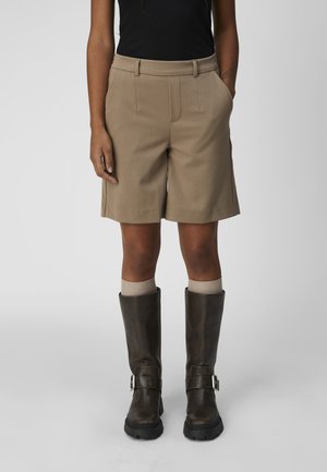 Personne portant un short beige jusqu'aux genoux, un haut noir sans manches, des chaussettes beiges et des bottines marron foncé à boucle, debout devant un fond uni.