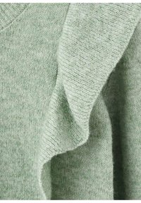 Svetlo zelena jopica z mehko, teksturirano tkanino. Ima v-izrez in volanasto podrobnost ob eni izmed rokavov.