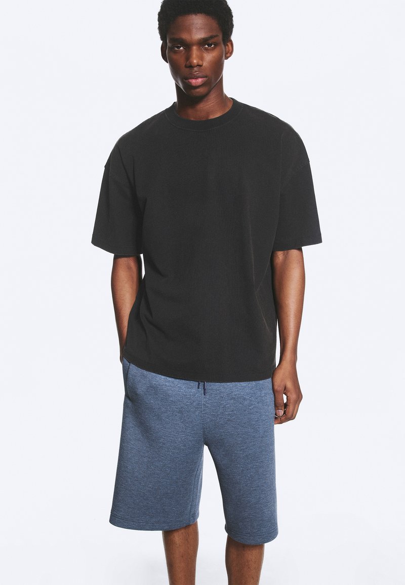 Jeune homme debout les mains dans les poches, portant un T-shirt noir oversize et un short bleu jusqu'aux genoux sur un fond uni.