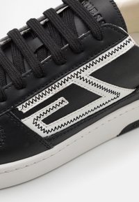 Baskets en cuir noir avec des lacets noirs, une couture en zigzag blanche et un logo blanc proéminent. Elles présentent une texture lisse et une semelle en caoutchouc.