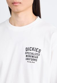 Tricou alb din bumbac cu gât rotund. Pe față are text negru: "DICKIES SPECIALIZED WORKWEAR UNIFORMS" și "Calitate Fabricată."
