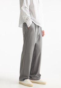 Pantalones grises con pliegues y un ajuste relajado, hechos de tela suave. Combinados con una camisa blanca a rayas y zapatillas de color claro.
