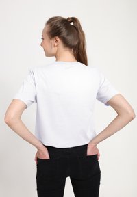 Bílé bavlněné tričko s krátkými rukávy, kulatým výstřihem, hladkou texturou, pohled zezadu, spojené s černými džínami s kapsami na zadní straně.