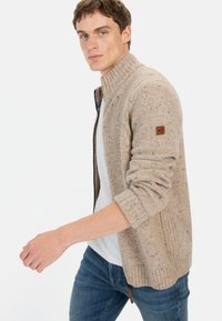 Beżowy sweter z dzianiny z wysokim kołnierzem, prążkowanymi mankietami i skórzaną naszywką z logo. Nosi się go na białej koszuli, w zestawieniu z niebieskimi dżinsami.