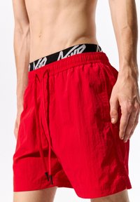 Pantaloni da bagno rossi con una cintura elastica e una fascia con logo nera. Tessuto texturizzato con una vestibilità rilassata e cordino regolabile.