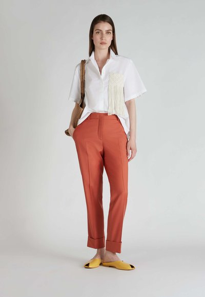 Camicia bianca a maniche corte con tasca in macramè texturizzato, abbinata a pantaloni su misura color ruggine e scarpe gialle slip-on.