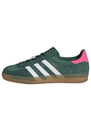 Groene suède Adidas sneaker met witte strepen, transparante gumzool en felroze hielstuk, zijaanzicht op witte achtergrond.