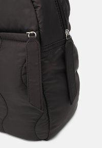 Černý polstrovaný batoh s texturovaným povrchem, zipovým uzavíráním a dvěma prodlouženými plochými popruhy v přední části pro zvýraznění detailu.