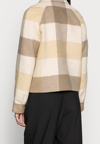 Villatakki, jossa on ruutukuvio beigen, kerman ja vaaleanruskean sävyissä. Takissa on kaulus, pitkät hihat ja lyhyt helma, jossa pehmeä, teksturoitu viimeistely.