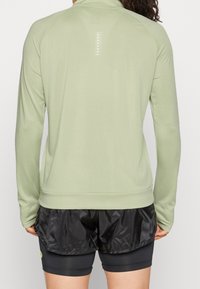 Ljusgrön långärmad sporttröja med hög krage, mjuk textur och reflektor detaljer längs ryggen. Matchas med svarta shorts.