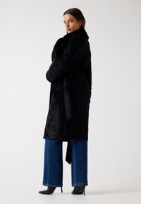 Schwarzer, strukturierter langer Mantel mit Gürtel, ausgestattet mit einem Zopfmuster und weichem Material, kombiniert mit weit geschnittenen blauen Jeans und schwarzen Absätzen.