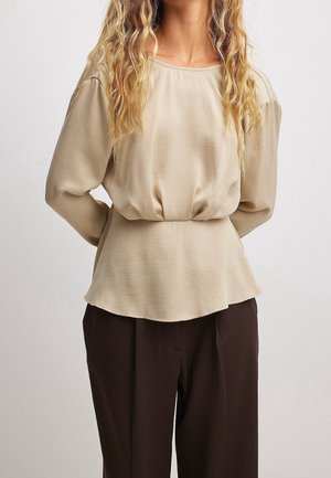 Mujer con cabello largo y ondulado rubio, que lleva una blusa beige de manga larga metida en pantalones marrones oscuros, de pie con las manos detrás de la espalda.