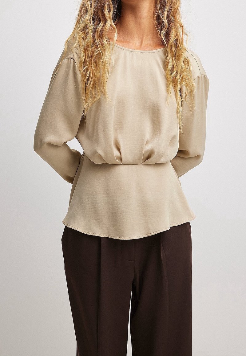 Femme aux longs cheveux blonds ondulés portant un chemisier beige à manches longues rentré dans un pantalon marron foncé, debout les mains derrière le dos.