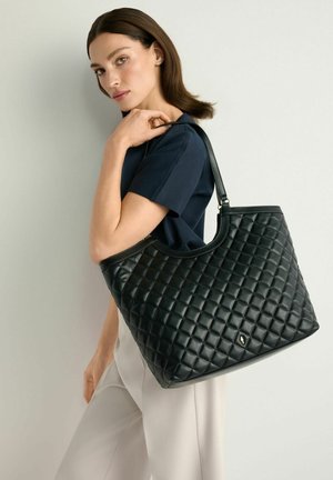 Kobieta o brązowych włosach do ramion ubrana w granatową bluzkę i białe spodnie, niesie na ramieniu dużą czarną pikowaną torebkę ze skóry.