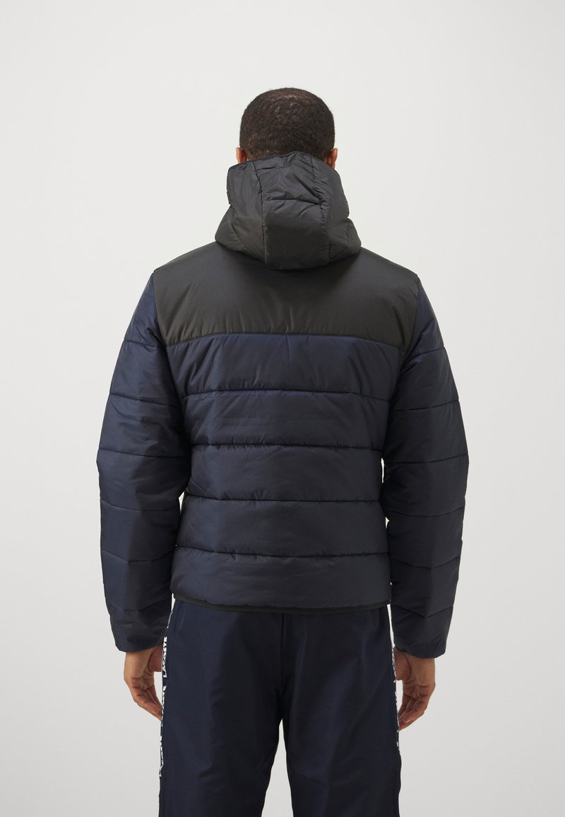Marineblaue Daunenjacke mit strukturiertem, gestepptem Design. Verfügt über ein schwarzes Paneel an den Schultern und eine verstellbare Kapuze. Schmale Passform mit elastischen Bündchen.