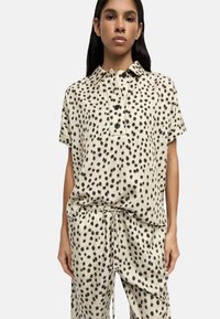 Krótki rękaw, bluzka z kołnierzykiem i spodnie w jasnym beżu z czarnymi plamkami, wykonane z miękkiego materiału; posiadają zapięcie na guziki z przodu oraz elastyczny pas.
