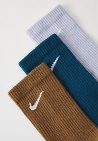 Three pairs of Nike socks: brown, teal, and white. Each features a ribbed texture and a white Nike swoosh logo on the front.