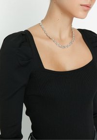 Zwarte geribbelde top met lange mouwen, een vierkante halslijn en pofmouwen, gecombineerd met een zilveren ketting met ronde schakels.