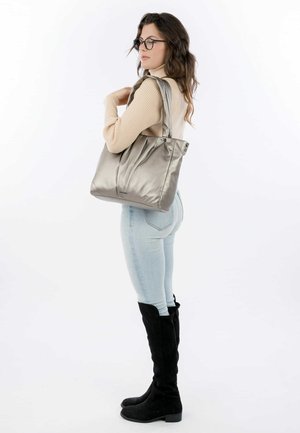 Silver tote bag with soft, shiny material. Features two short handles and gathered details. Worn with light blue jeans and black knee-high boots.