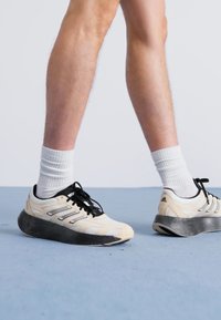 Baskets de sport beiges et noires avec une semelle en caoutchouc texturée, dotées de panneaux en mesh blanc et de lacets noirs. Portées avec des chaussettes blanches.
