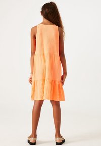 Vestido naranja de volantes con un acabado texturizado, diseño sin mangas y parte inferior acampanada, combinado con sandalias planas.