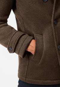Brown textured jacket with a close-fitting collar, button details on cuffs, and side pockets. Fabric has a woven pattern and sturdy construction.