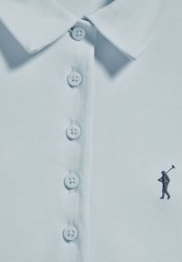 Polo bleu clair en tissu texturé, avec un col classique, cinq boutons et un petit logo brodé en bas à droite.