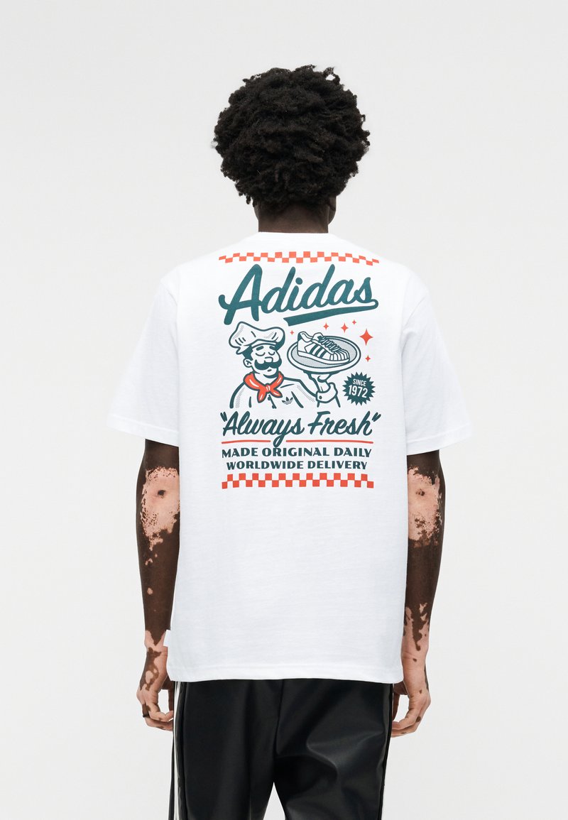 Sötét, göndör hajú személy, aki fehér Adidas pólót visel egy retró séf és sportcipő grafikával, valamint "Always Fresh" felirattal a hátoldalán.