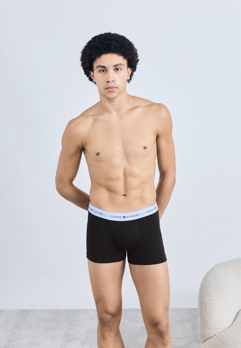 Mustad bokserid valge vöökohaga, millel on "TOMMY HILFIGER" logo, esitletud meessoost modellil neutraalsel taustal.