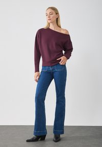 Sudadera burdeos de hombros descubiertos hecha de una tela suave, combinada con jeans acampanados azules y botas negras de tacón, contra un fondo plano.