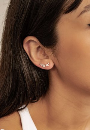 Minimalistische Silberohrringe mit fünf kleinen klaren Steinen, die in einer gebogenen Linie angeordnet sind, vor dem Ohr eines Models und dunklem Haar.